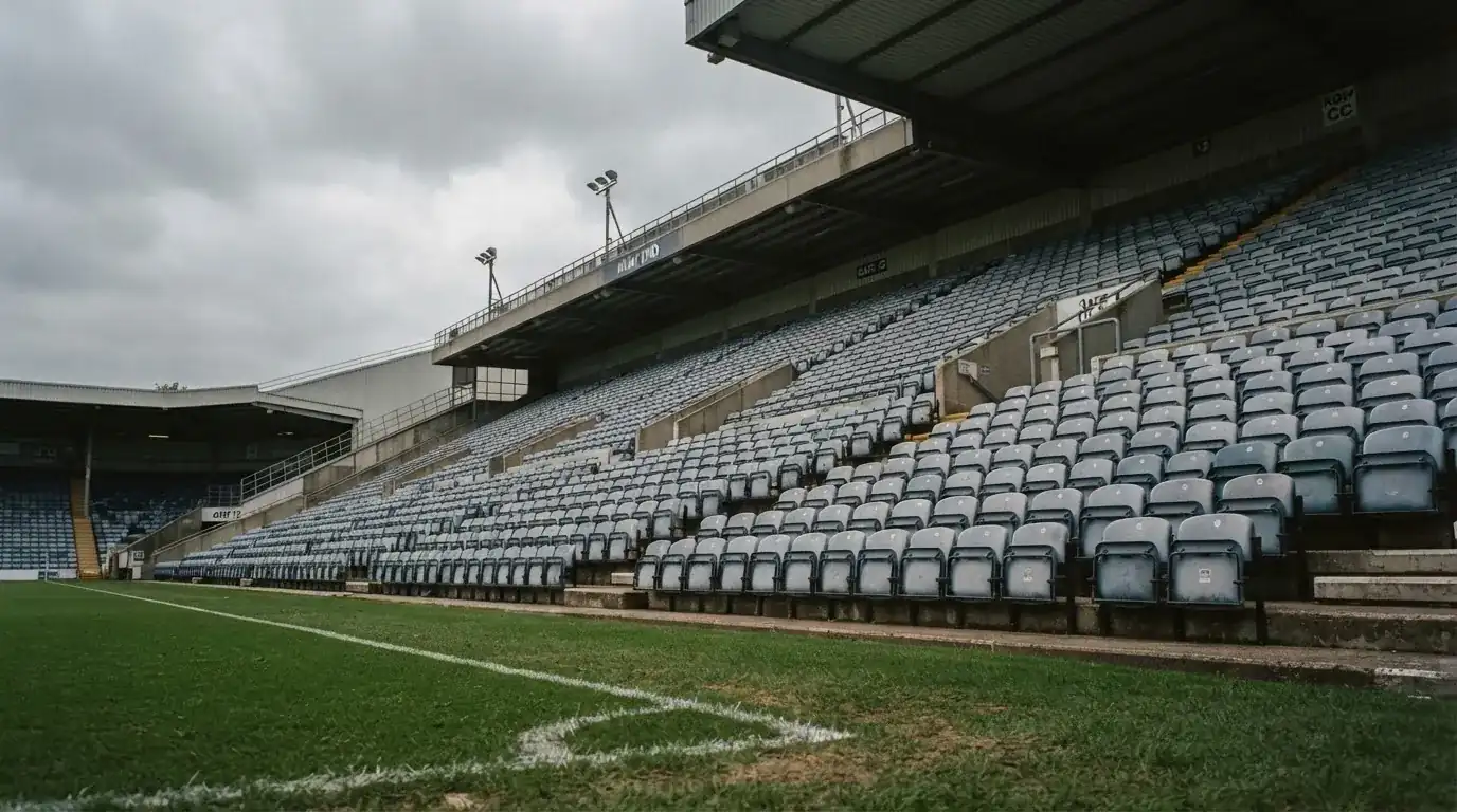 Rzędy pustych plastikowych krzesełek małego stadionu widziane z poziomu murawy pod pochmurnym niebem.