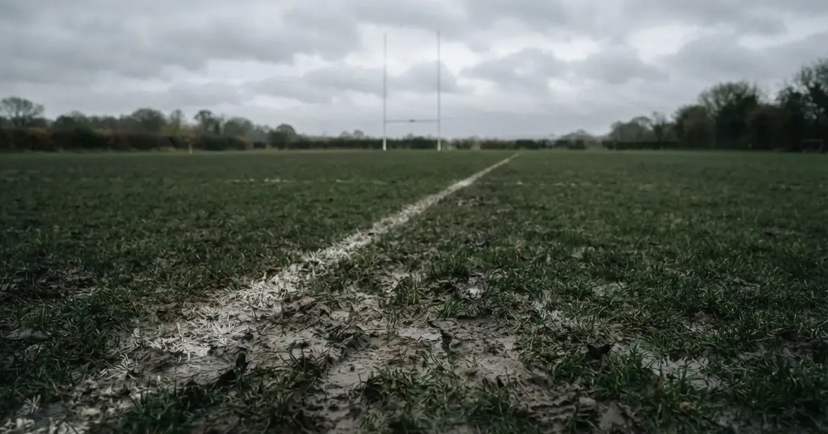 Boisko do rugby ze słupami bramki w oddali pod pochmurnym niebem, mokra trawa i ślady butów.