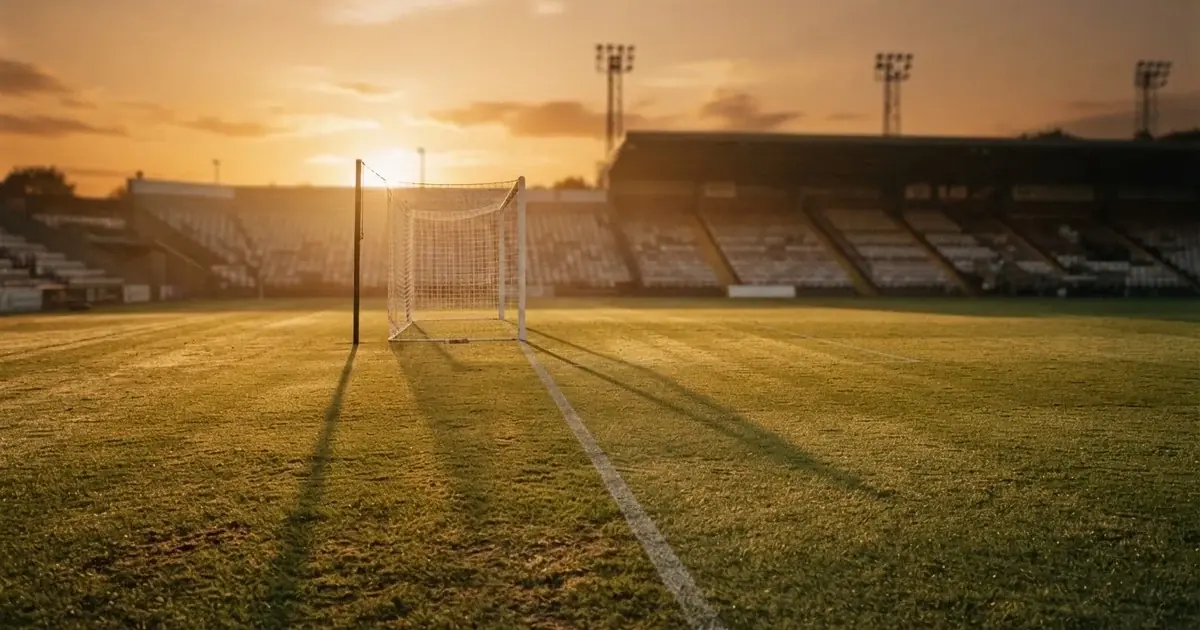 Murawa stadionu o zachodzie słońca z długimi cieniami słupków bramki, trybuny w ciepłym świetle.